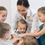 "Enseignante souriante mettant ses mains en cercle avec un groupe d'enfants jouant à un jeu."