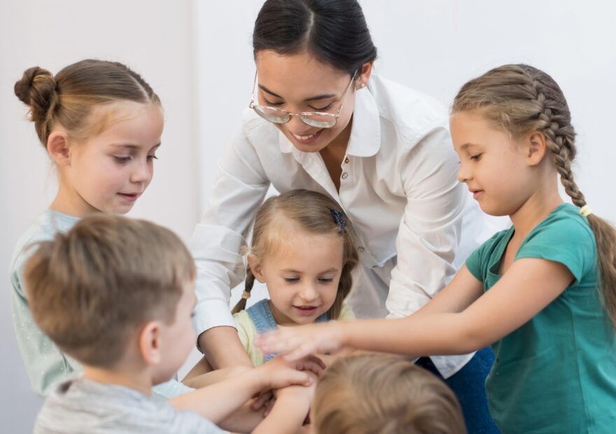 "Enseignante souriante mettant ses mains en cercle avec un groupe d'enfants jouant à un jeu."